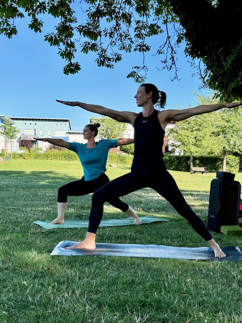 Isabel Stapf und Alix Steinhoff praktizieren im Freien die Yoga-Haltung Krieger II während eines -Outdoor-Yoga-Kurses im Schlosspark Butzbach unter blauem Himmel.